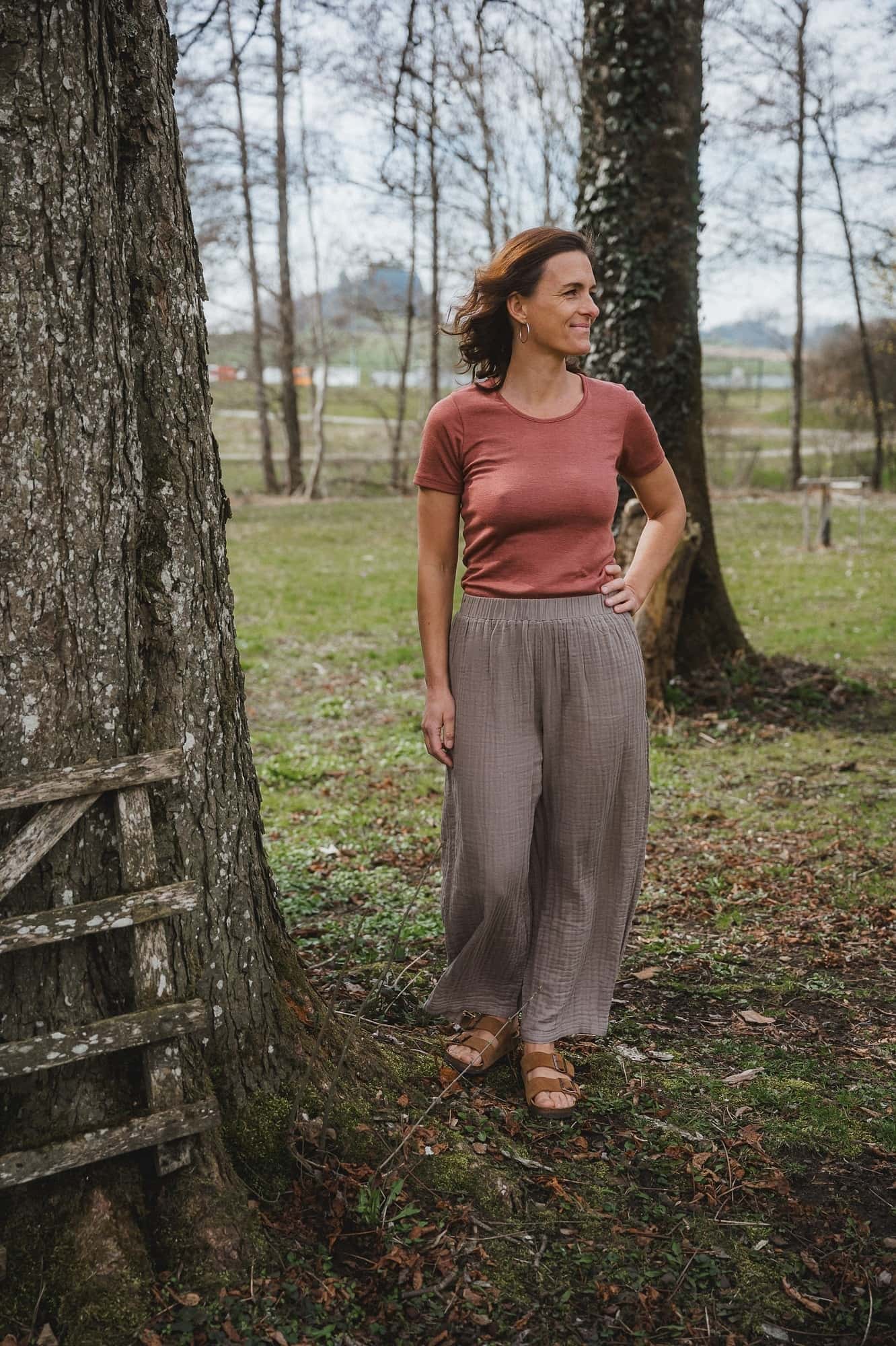 Damenhose aus Bio-Baumwoll-Musselin Frau steht neben Baum und trägt Shirt und braune weite Hose aus Bio-Baumwoll-Musselin vom Finkhof