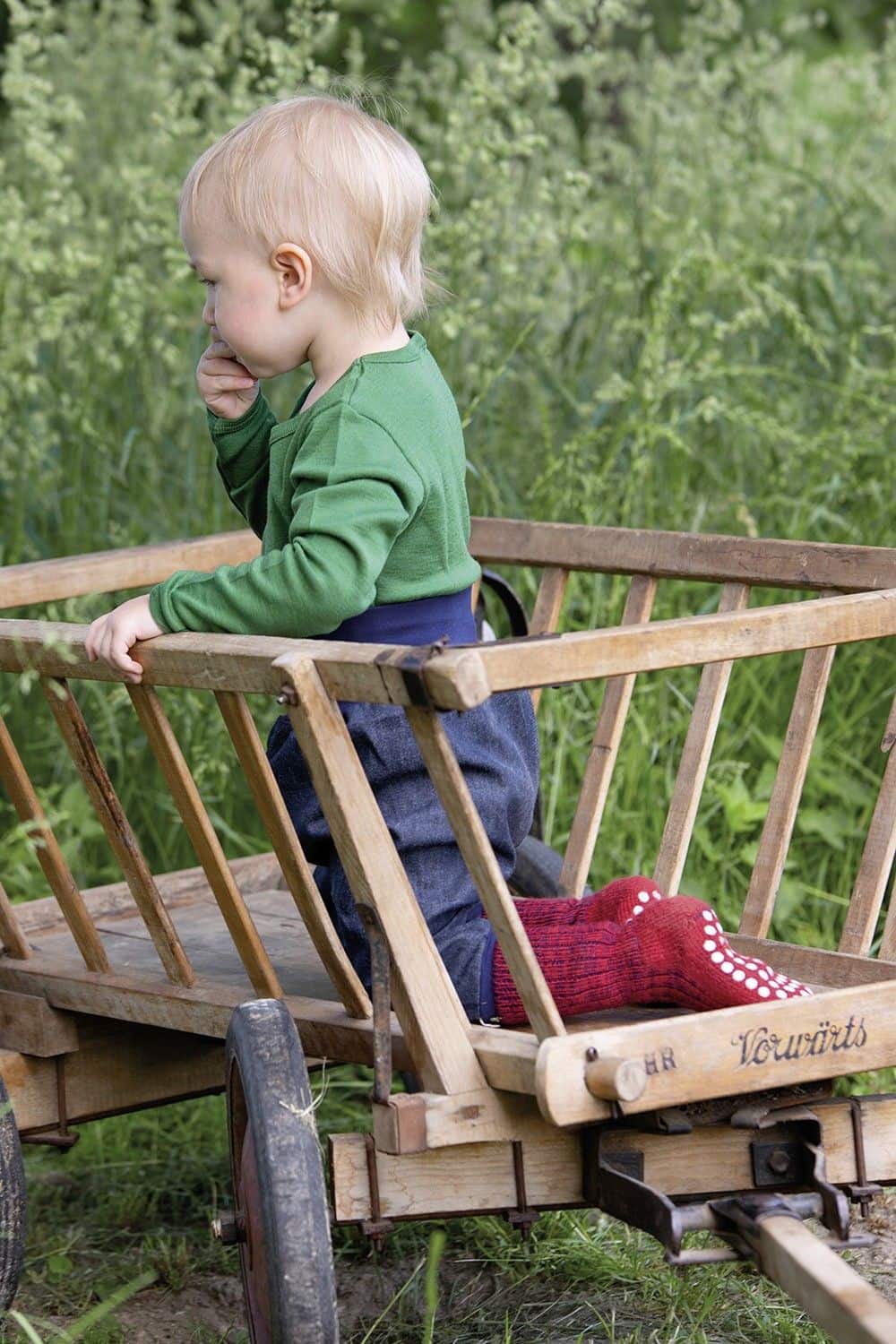 Kind in Leiterwagen trägt blaue Hose, grünes Shirt und rote Wollplüsch Stoppersocken aus bio Wolle (kbT)