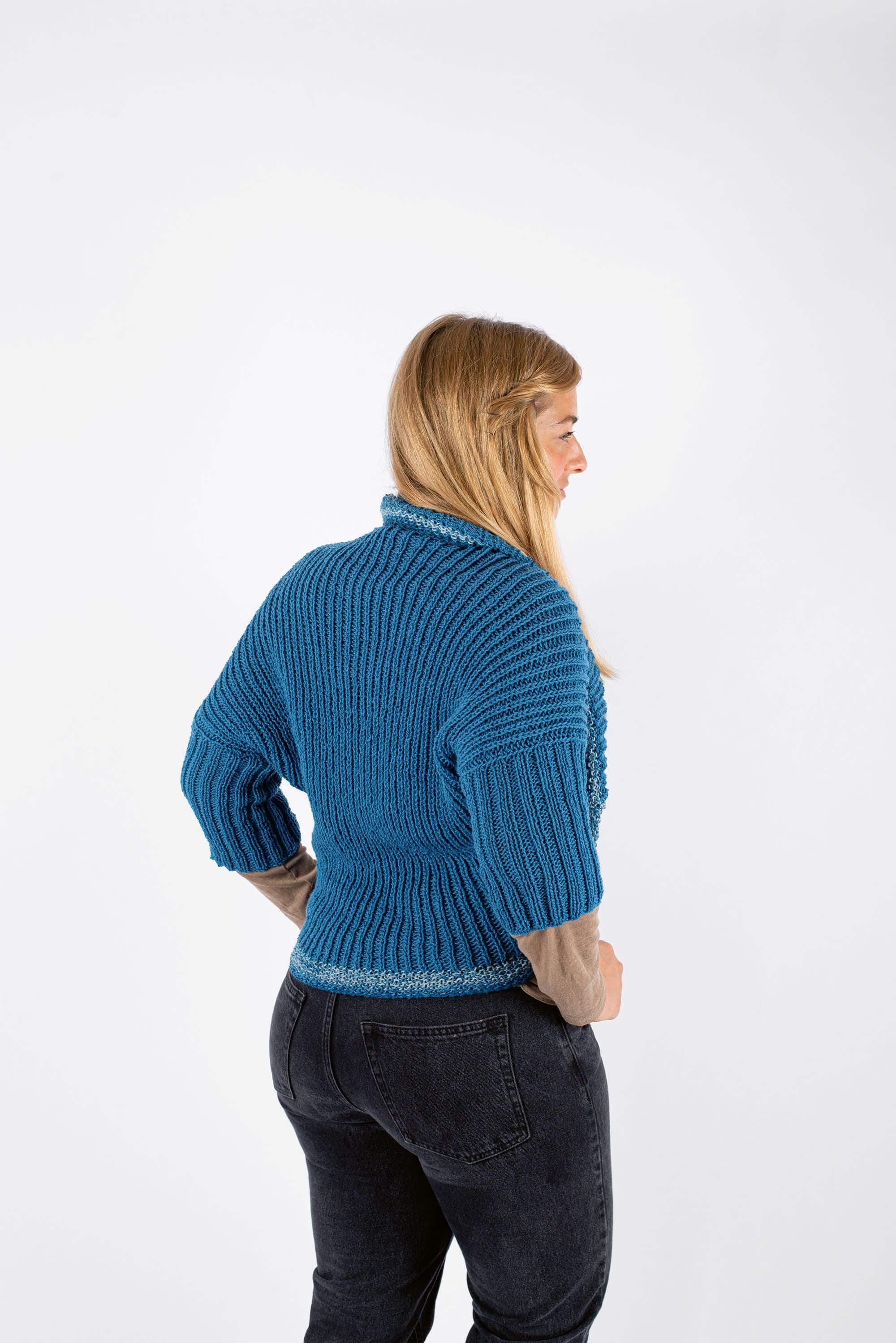 Frau mit blonden Haaren hat sich blauen Strickseelenwärmer um Schultern gelegt, von hinten aufgenommen