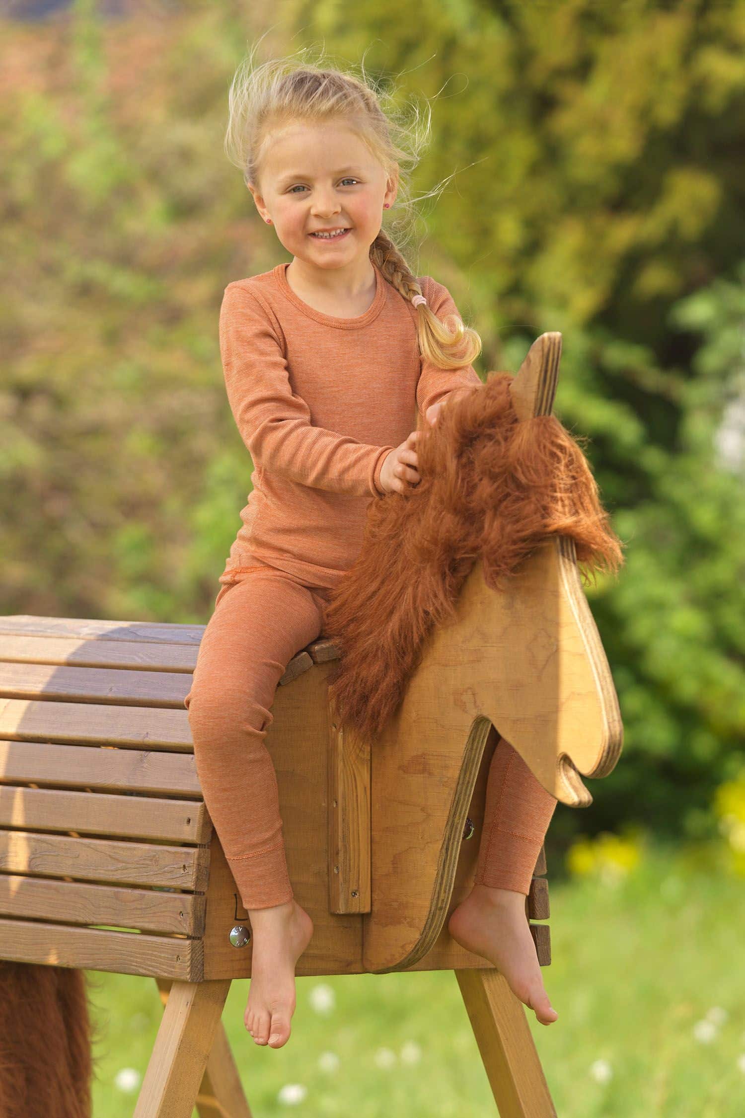 Kind sitzt auf Holzpferd und trägt ein oranges Langarmshirt aus bio Wolle (kbT), bio Baumwolle (kbA) und Seide mit Hose