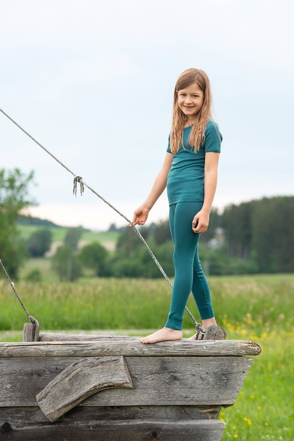Kind spielt auf Holzschiff und trägt türkise lange Unterhose und Shirt aus bio Wolle (kbT) und Seide