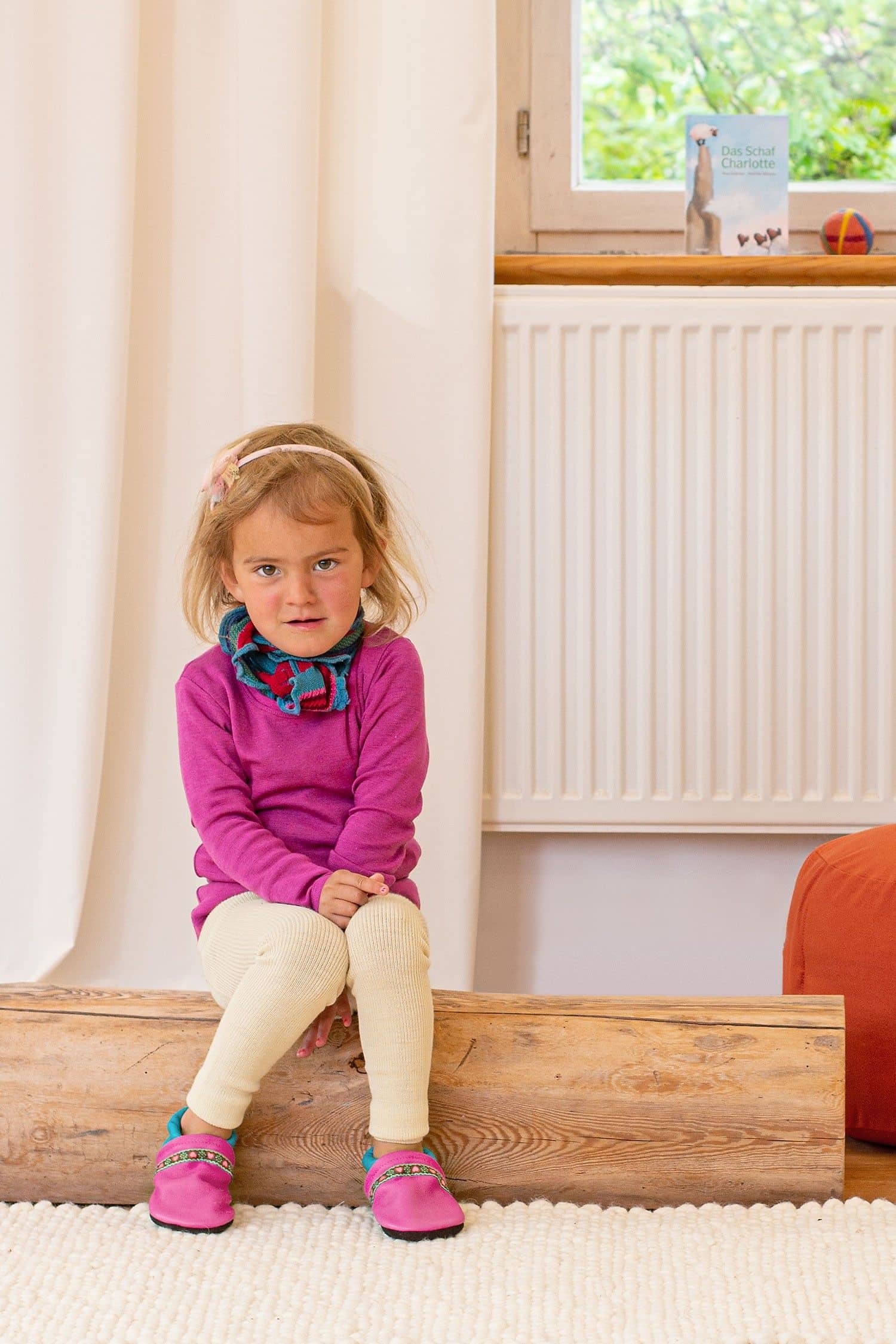Kind sitzt auf Stamm und trägt pinkes Shirt, bunten Schal und naturweiße Woll-Leggings aus bio Wolle (kbT)
