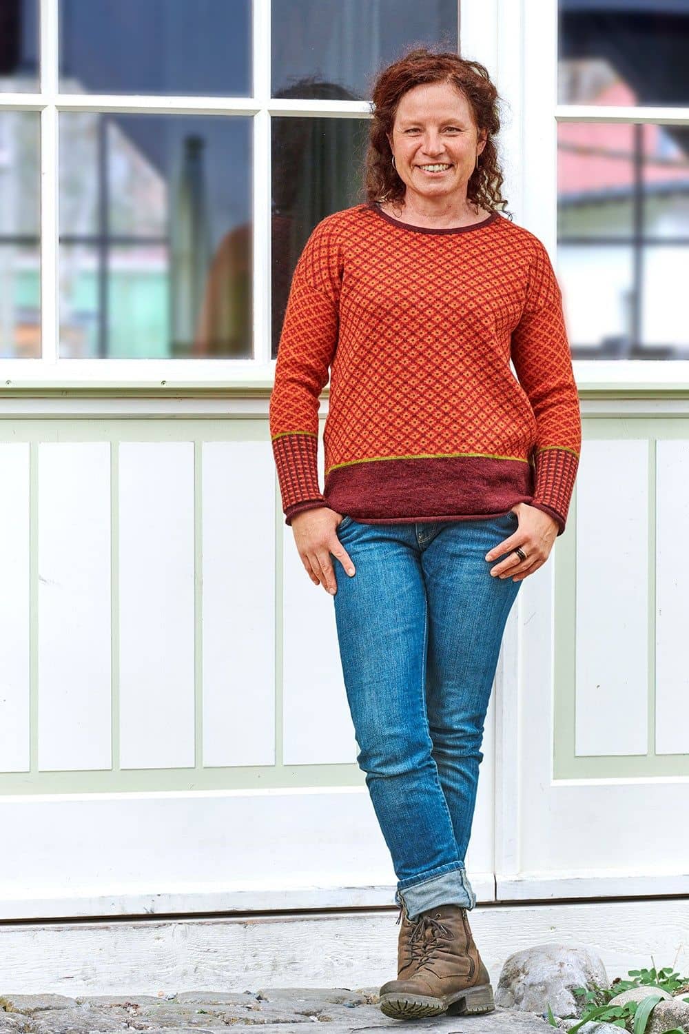 Frau steht vor Gartenhütte und trägt rot orange gemusterten Pulli aus bio Wolle (kbT) mit Jeans