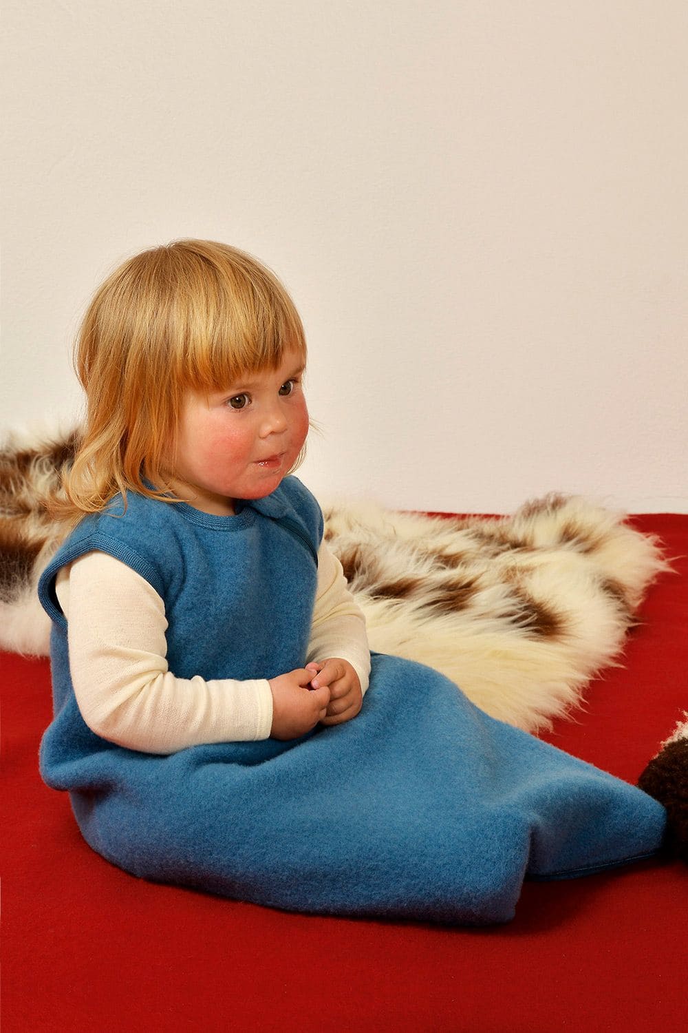 Wollfleece-Schlafsack blau Kind trägt blauer Wollfleece Schlafsack aus bio Wolle (kbT) und sitzt auf rotem Laken mit Fell im Hintergrund