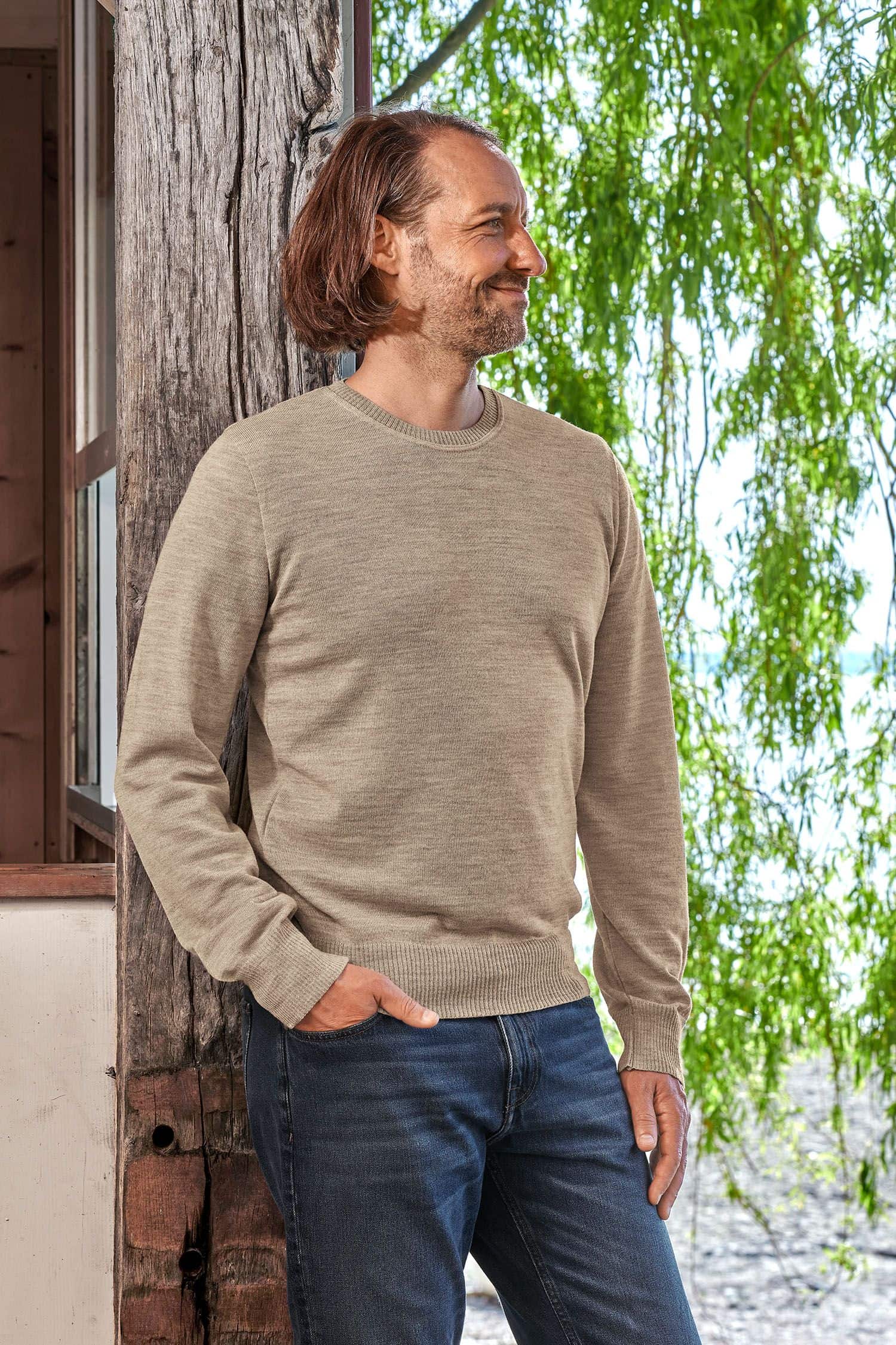 Mann mit feinem beigen Wollpullover aus bio Merinowolle (kbT) vom Finkhof lehnt an einem Balken