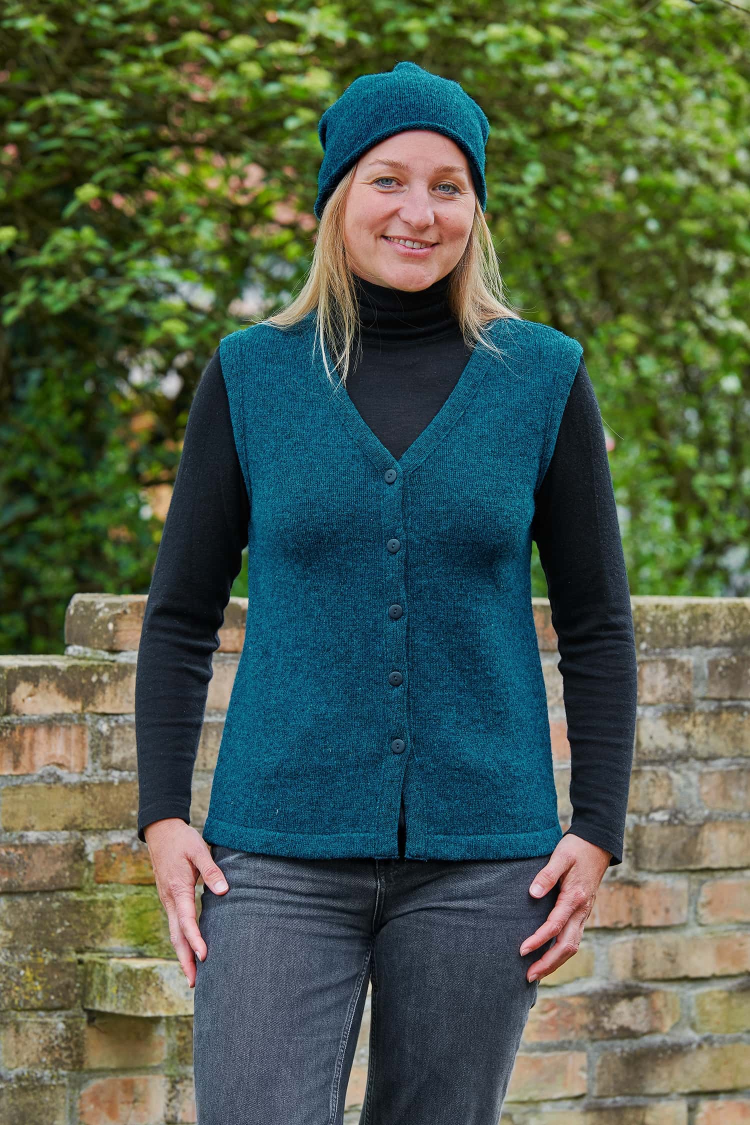Frau mit Jeans und schwarzem Shirt trägt eine türkise Mütze und eine feine türkise Strickweste vom Finkhof aus Bio-Wolle