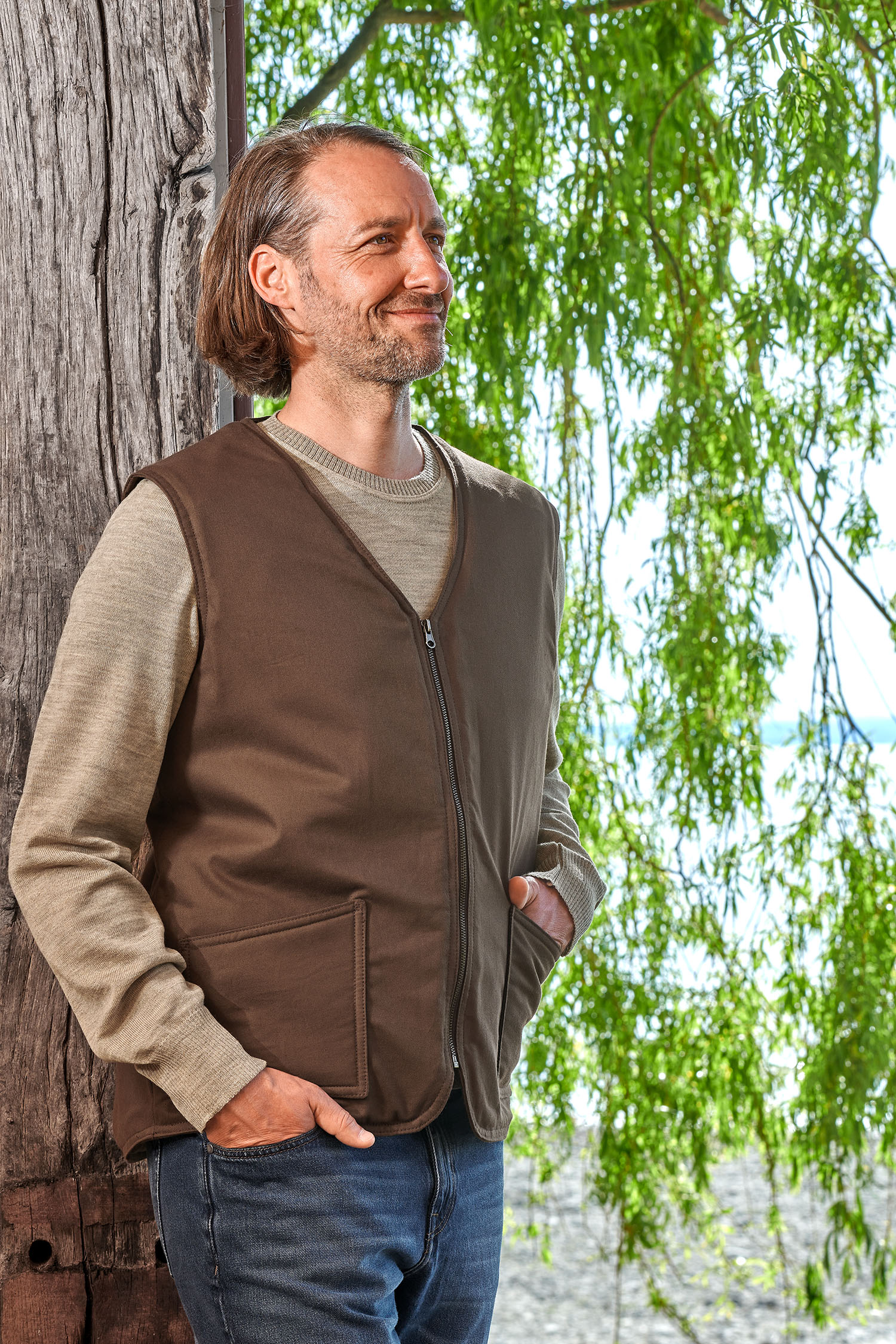 Mann lehnt sich an Baum und trägt die braune Fellweste aus bio Baumwolle (kbA) und Fell vom Finkhof