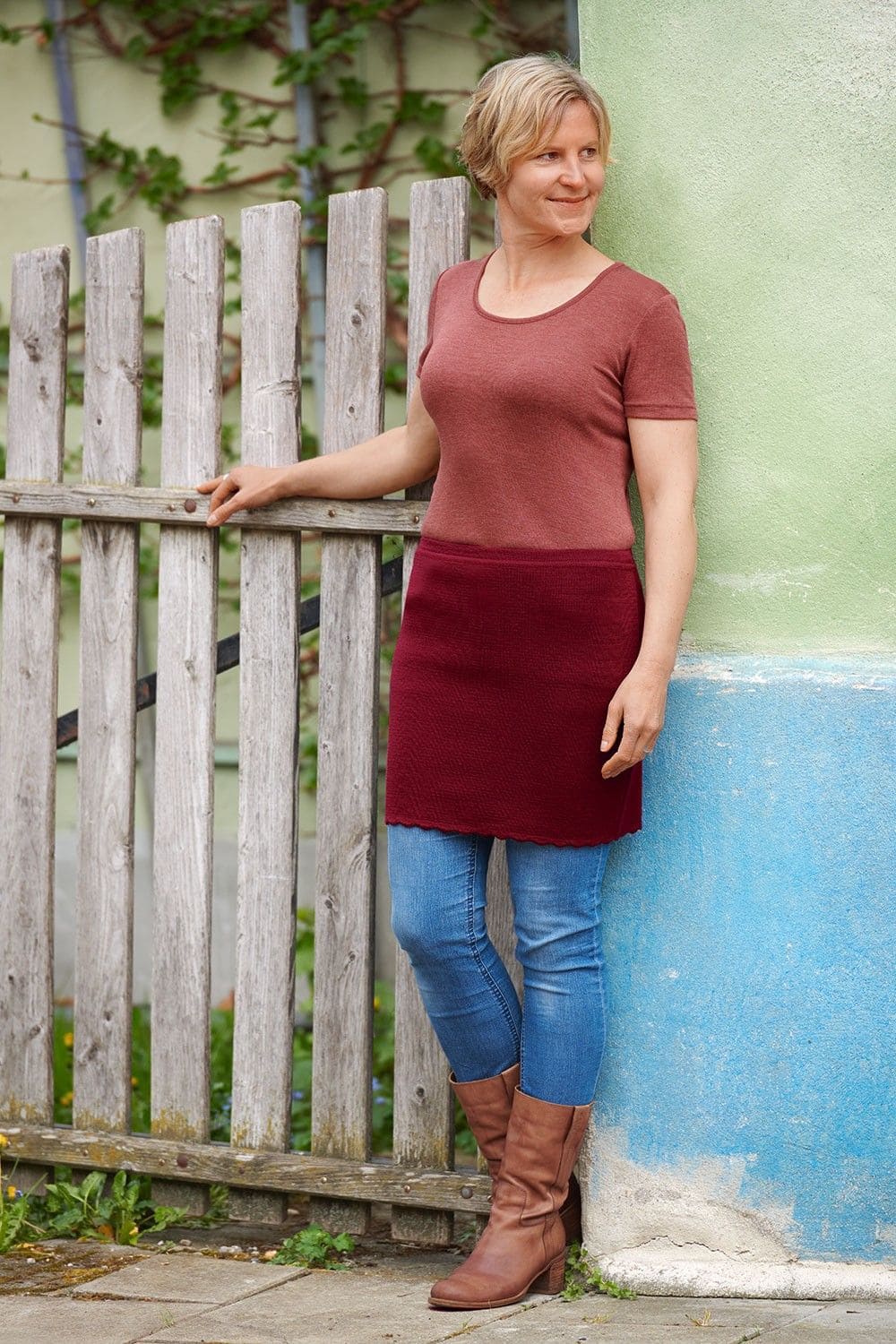 Frau mit rotem Shirt und rotem Kurzrock aus bio Wolle (kbT) vom Finkhof lehnt an grüner Hauswand